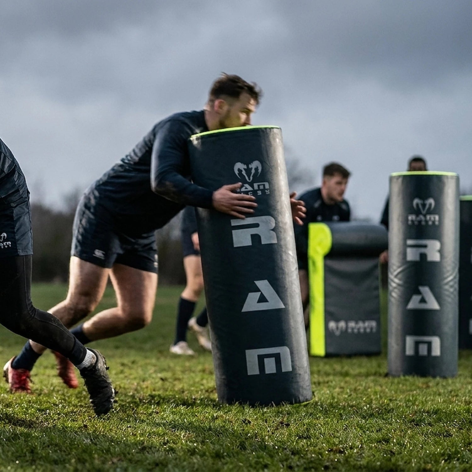 Athletes training with black and neon yellow rugby scrum pads on a grassy field under an overcast sky. The pads feature the RAM Rugby logo.
